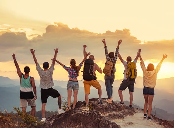 friends jump and raise their hands with their backs to the camera, while clouds obscure the sunset